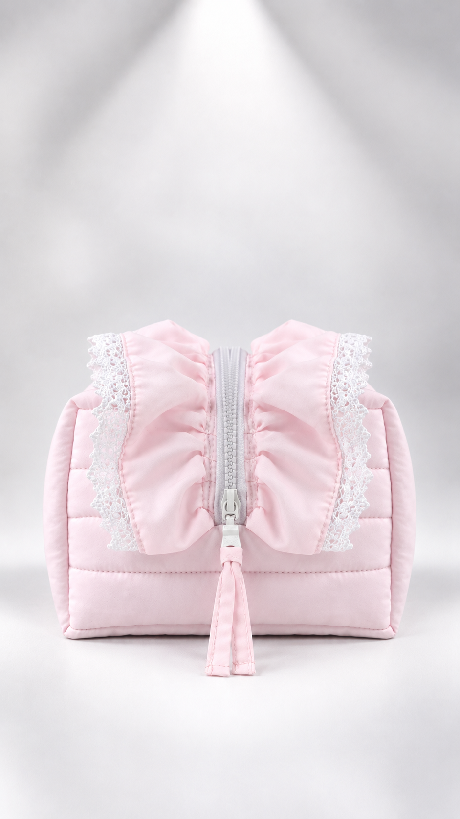 A light pink quilted cosmetic bag with a large bow on the front, white lace trim, a silver zipper, and a matching pink tassel, set against a soft gray studio background.