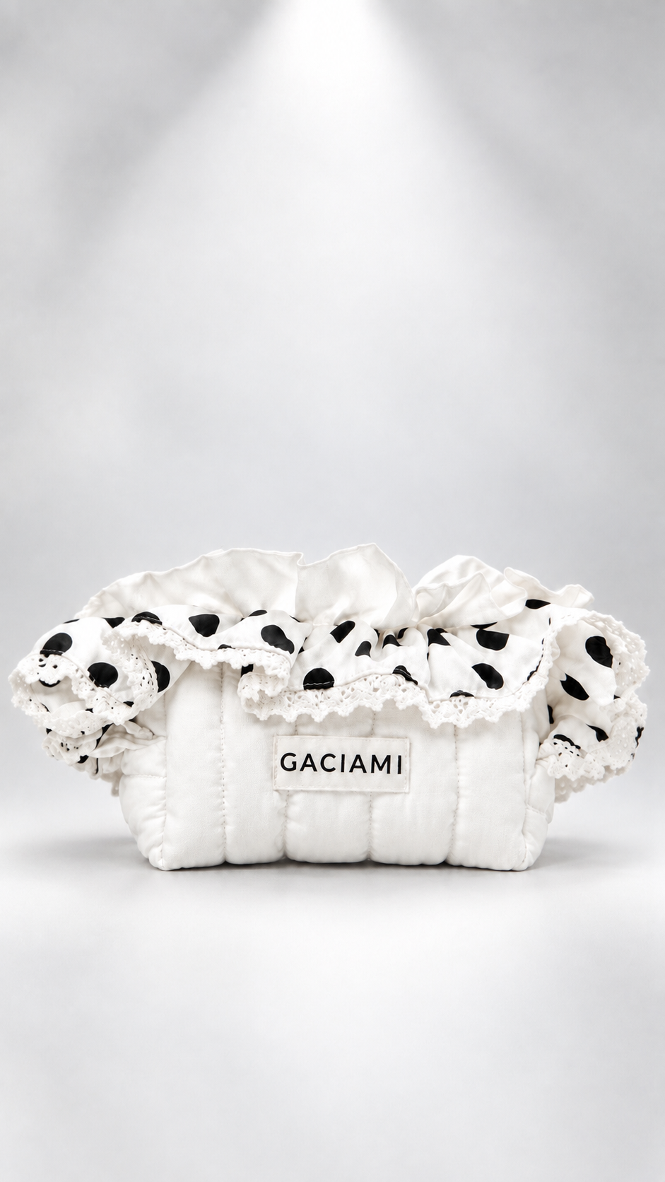A white quilted clutch bag with black polka-dot lace trim and a white label reading 'GACIAMI', photographed against a soft gray studio background with a spotlight effect.