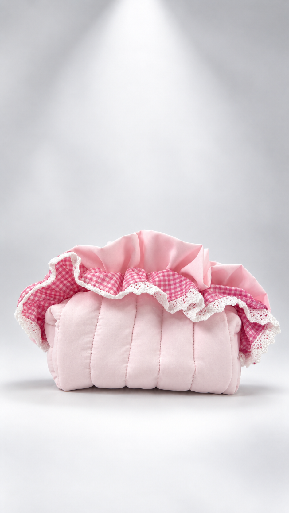 A pink quilted cushion cover with ruffled trim, featuring a pink and white gingham pattern and white lace trim, set against a light gray background with a spotlight effect.