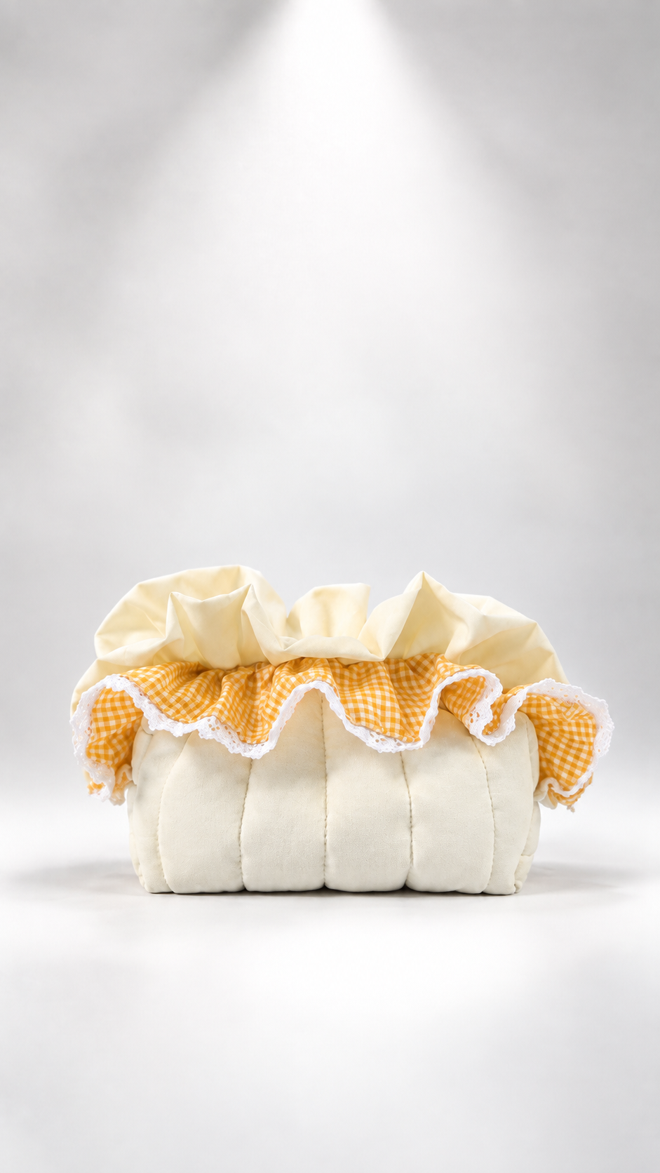 A cream-colored quilted fabric pouf or cushion with a ruffled top trim in yellow and white gingham and white lace, set against a light gray background with a spotlight effect.