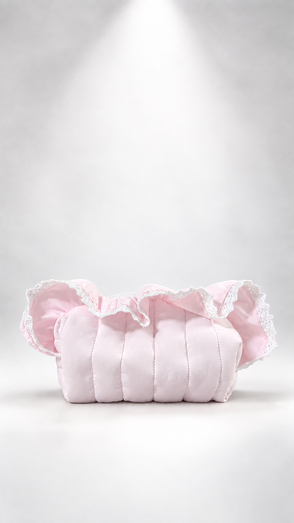 A light pink quilted fabric pouch or small bag with ruffled trim, photographed against a soft white studio background with a spotlight effect above.