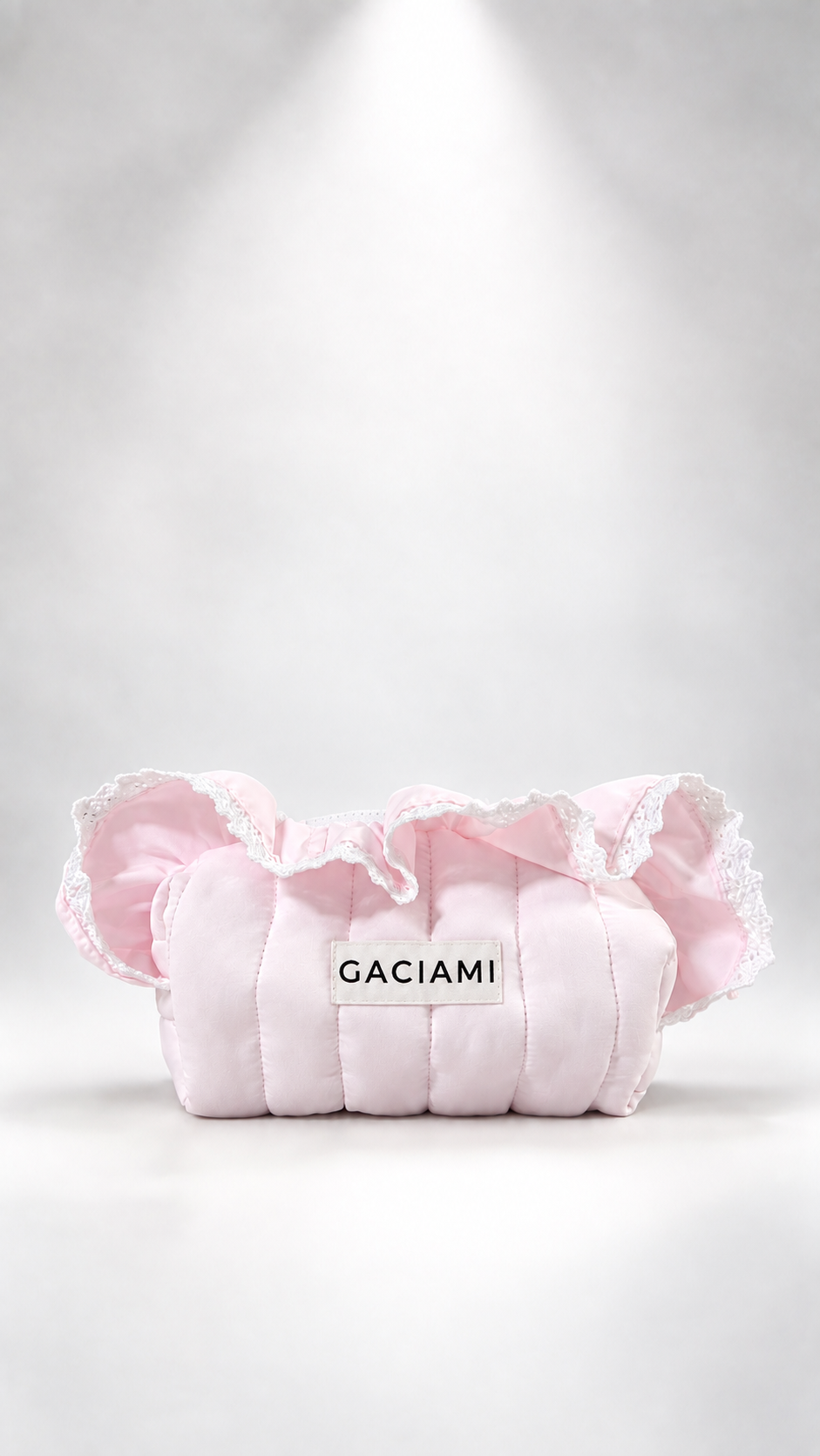 A light pink quilted cosmetic pouch with ruffled trim and a white label reading 'GACIAMI' in black text, photographed on a white studio background with a spotlight effect.