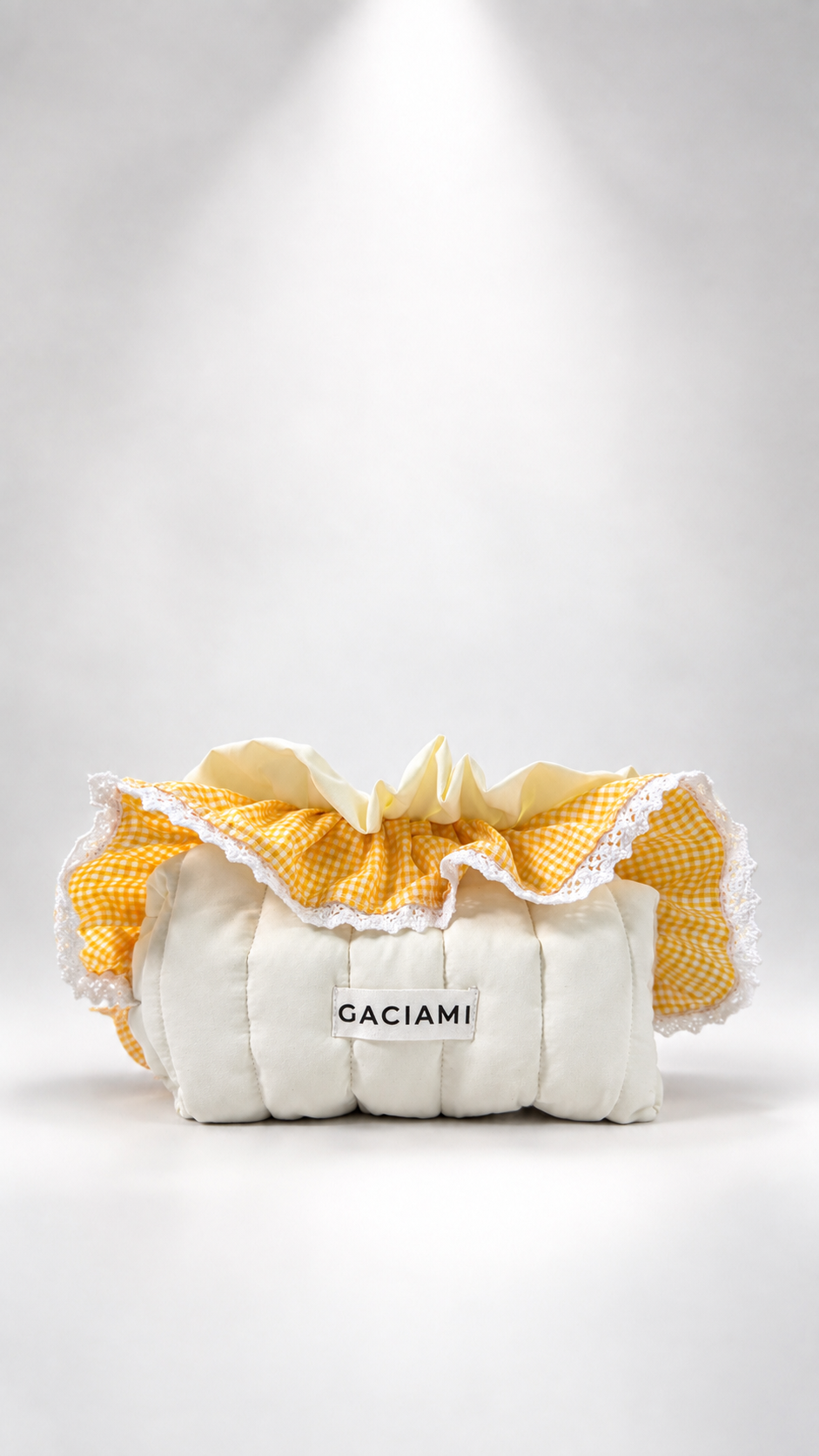 A cream-colored quilted clutch bag with a yellow and white gingham ruffled fabric top trim, featuring a white label with the brand name 'GACIAMI' in black text, set against a white studio background with a spotlight effect.