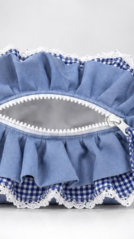 A close-up of a blue ruffled fabric pouch with a white zipper, white lace trim, and a blue-and-white gingham patterned lining, shown against a light gray background.