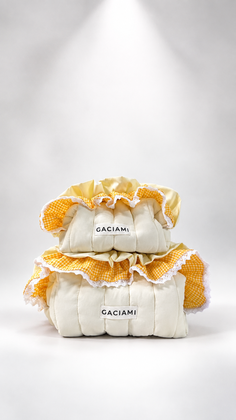 Three stacked quilted cream-colored pillows with yellow and white gingham ruffled trim, featuring the brand name 'GACIAMI' on the front labels, set against a white studio background with a spotlight effect.